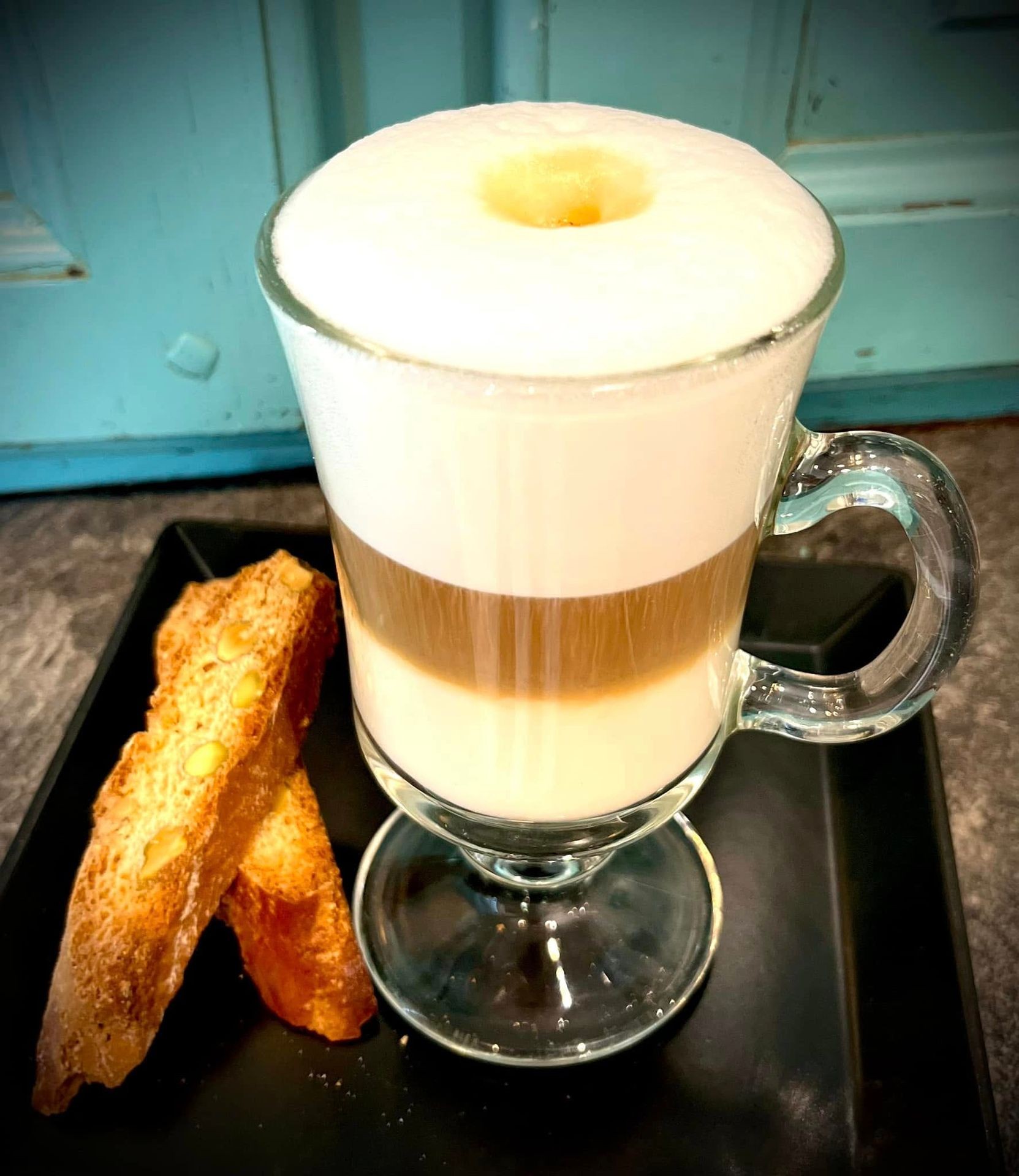 Glass of cappuccino with frothy milk and biscotti on a black tray.