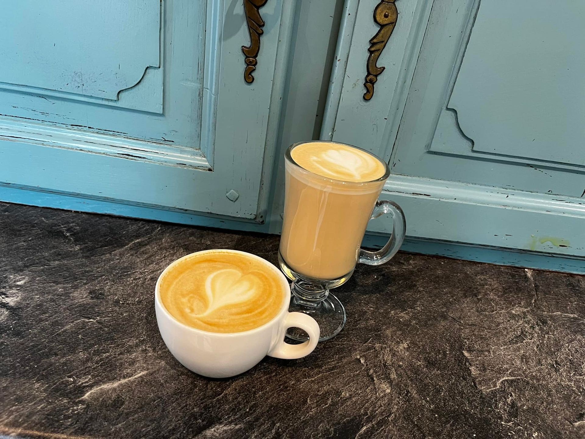 Two cups of latte with heart-shaped latte art on a dark marble countertop and blue cabinet background.