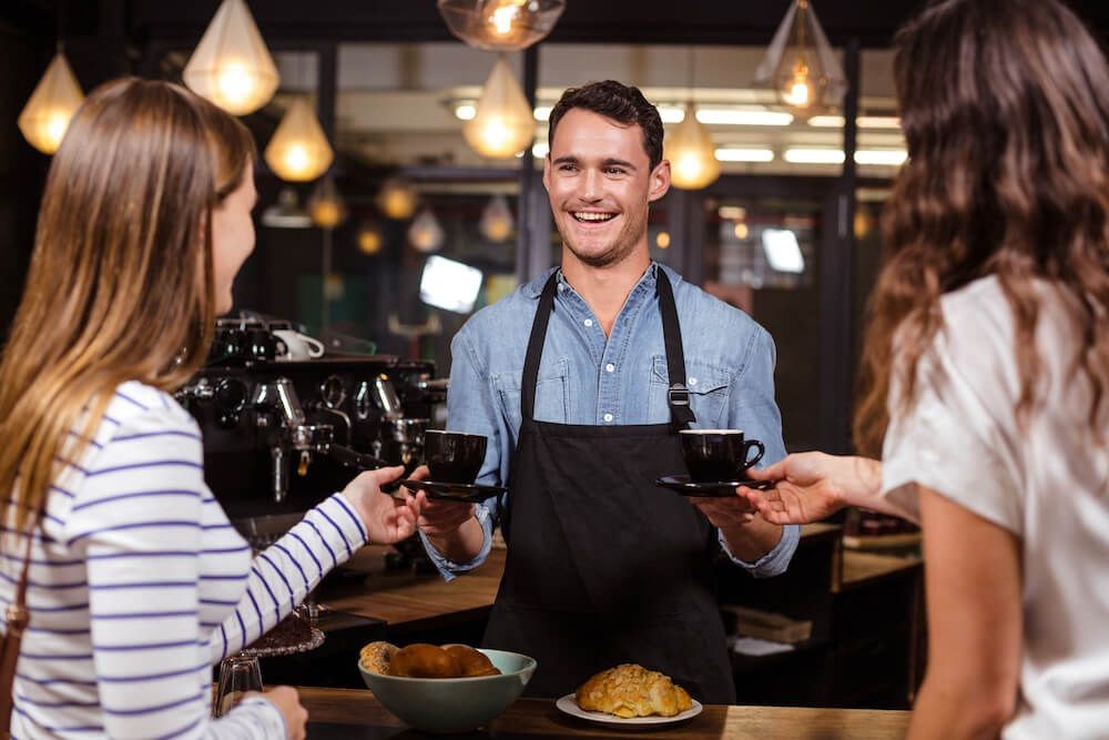 Smiling barista serving coffee cups to two women in a cozy cafe setting with warm lighting.